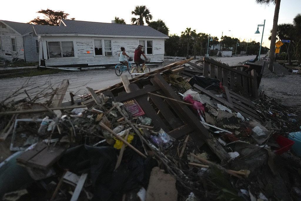 Personas andan en bicicleta frente a escombros y viviendas dañadas por el paso del huracán Milton en la calle principal de Manasota Key, el domingo 13 de octubre de 2024, en Englewood, Florida. (AP Foto/Rebecca Blackwell)