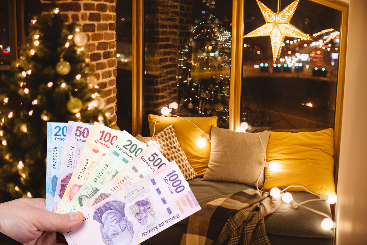 De fondo casa adornada con Navidad, mano con billetes de aguinaldo. Foto: Canva