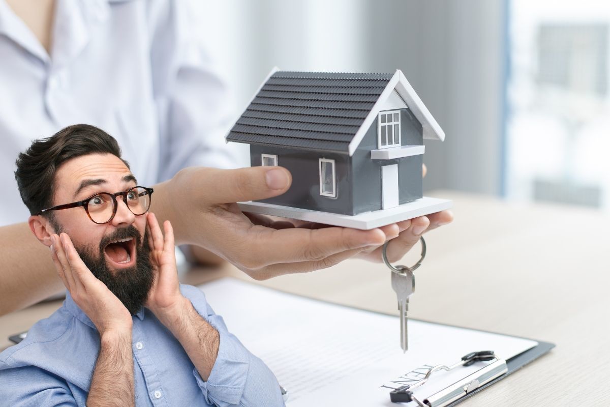 Persona sosteniendo una casa a escala junto con unas llaves y en la parte inferior izquierda, un hombre sorprendido.    Foto: Canva